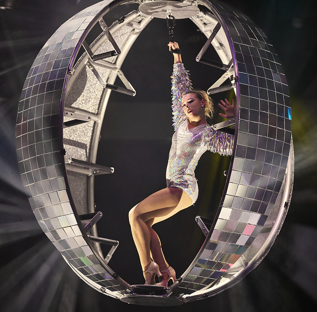 An acrobat in a silver metallic suit performs on a sphere resembling a disco ball, suspended in the air